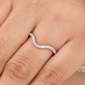 Pode incluir: Um anel prateado curvo com pequenas pedras preciosas transparentes. O anel tem um design em forma de onda, com as pedras preciosas seguindo a curva. O anel é mostrado em um dedo.