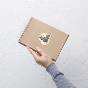Puede incluir: Una pegatina con una bruja de dibujos animados con sombrero y capa negros, sosteniendo una varita m&aacute;gica. La bruja est&aacute; rodeada de un dise&ntilde;o floral amarillo y texto. La pegatina est&aacute; sobre un cuaderno en espiral marr&oacute;n.