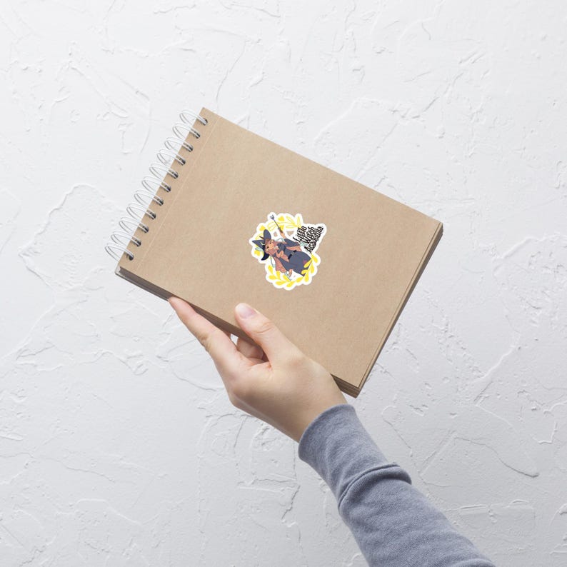 Puede incluir: Una pegatina con una bruja de dibujos animados con sombrero y capa negros, sosteniendo una varita m&aacute;gica. La bruja est&aacute; rodeada de un dise&ntilde;o floral amarillo y texto. La pegatina est&aacute; sobre un cuaderno en espiral marr&oacute;n.