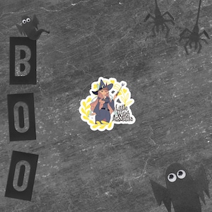 Puede incluir: Una pegatina con una bruja de dibujos animados con pelo rosa, un atuendo azul y una varita m&aacute;gica, rodeada de acentos florales amarillos. La pegatina est&aacute; etiquetada como "Little Witch Academia". La palabra "BOO" est&aacute; escrita con letras negras, y hay recortes de papel de ara&ntilde;as y fantasmas.
