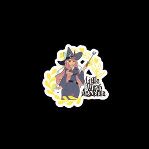 Puede incluir: Una pegatina con una ilustraci&oacute;n de estilo anime de una bruja con pelo rosa, vestida con un atuendo azul oscuro y un sombrero puntiagudo con una luna creciente. El texto "Little Witch Academia" tambi&eacute;n est&aacute; en la pegatina, rodeado de detalles florales amarillos.
