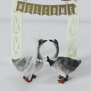 May include: A wedding cake topper featuring two goose figurines facing each other. One goose wears a veil and pearl necklace, the other a bow tie. A decorative arch with the word "WELCOME" is in the background, with floral and bird accents.
