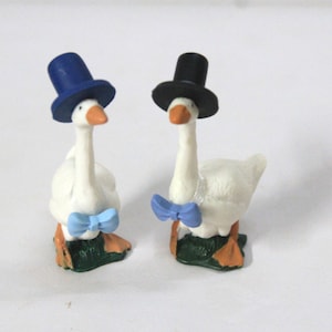 May include: Two white goose figurines standing on green bases. The left goose wears a blue top hat and a light blue bow tie. The right goose wears a black top hat and a light blue bow tie. Both geese have orange beaks and orange webbed feet.