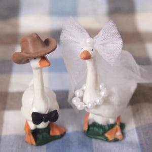 May include: Two ceramic goose figurines, one wearing a brown cowboy hat and a black bow tie, the other adorned with a white veil and pearl necklace. The geese have orange beaks and feet, standing on green bases.