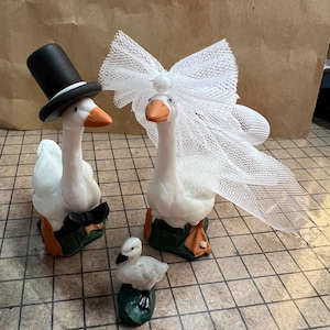 May include: Three ceramic goose figurines. One goose wears a black top hat and bow tie, another wears a white veil, and a smaller goose figurine is also present. The geese have white bodies, orange beaks and feet, and are on green bases.