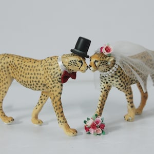 May include: Two cheetah figurines, one wearing a black top hat and bow tie, the other a veil and floral headpiece. They are positioned nose-to-nose, with a small bouquet of flowers at their feet. The cheetahs have a spotted pattern.