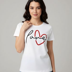 Puede incluir: Camiseta blanca con la palabra "Love" en escritura negra y un diseño de corazón rojo. La camiseta es de estilo cuello redondo clásico y parece estar hecha de un material suave.