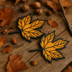 Puede incluir: Dos posavasos amarillos en forma de hoja de arce con contornos y tallos negros se muestran sobre una superficie de madera, rodeados de hojas caídas y bellotas. Los posavasos tienen una combinación de colores otoñal vibrante.