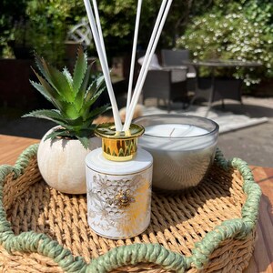 Könnte beinhalten: Ein dekoratives Tablett mit einem weißen und goldenen Blumendiffusor mit weißen Stäbchen, einer kleinen Sukkulente und einer Glaskerze. Das Tablett hat ein gewebtes Design mit grüner Seilborte. Auf einer Holzoberfläche angeordnet.