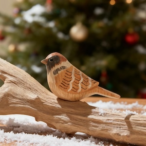 以下が含まれることがあります： 茶色と黒の模様が入った木製の鳥の置物が枝に止まっています。背景にはオーナメントが飾られたクリスマスツリーがあり、お祝いのシーンを演出しています。