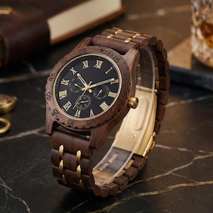 May include: A wooden wristwatch with a dark face and gold Roman numerals. The watch has a brown wooden band with gold accents. The watch is displayed on a dark surface, with a leather-bound book and a glass of amber liquid in the background.