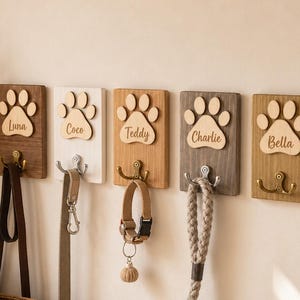 Op de afbeelding: Vijf rechthoekige houten wandhaken met uitgesneden pootafdrukken en de namen Luna, Coco, Teddy, Charlie en Bella. Elke plaat heeft een metalen haak voor diverse hondenriemen en halsbanden, gemonteerd in een rij op een lichte muur.