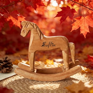 Könnte beinhalten: Ein Holzschaukelpferd mit dem eingravierten Namen "Sofia" auf der Seite. Das Pferd ist hellbraun und steht auf einer geschwungenen Basis. Der Hintergrund zeigt rote und orangefarbene Herbstblätter, die eine warme, saisonale Szene schaffen.