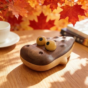 May include: A wooden alligator ornament with two yellow eyes with black pupils. The alligator is made of dark and light brown wood, with a textured back. The ornament is on a wooden surface, with autumn leaves in the background.