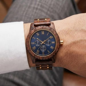 Peut inclure: Une montre-bracelet en bois avec un cadran bleu fonc&eacute; et des aiguilles et accents dor&eacute;s. La montre comporte des chiffres romains et trois sous-cadrans. Le bracelet et la lunette sont en bois. La montre est au poignet.