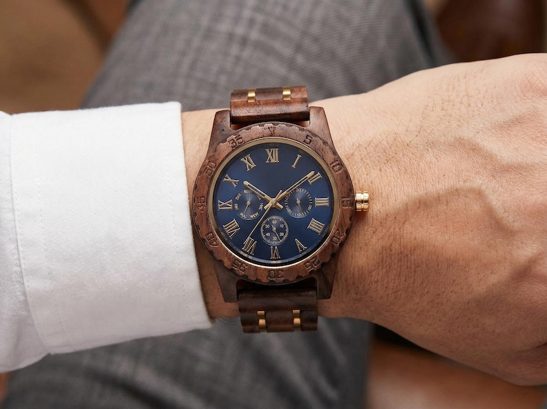 Peut inclure: Une montre-bracelet en bois avec un cadran bleu fonc&eacute; et des aiguilles et accents dor&eacute;s. La montre comporte des chiffres romains et trois sous-cadrans. Le bracelet et la lunette sont en bois. La montre est au poignet.