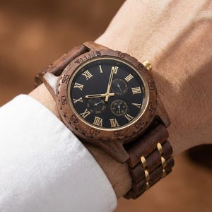 Peut inclure: Montre-bracelet en bois avec un cadran noir et des chiffres romains dor&eacute;s. La montre a une lunette et un bracelet en bois marron avec des accents dor&eacute;s. La montre est sur un poignet avec une manche de chemise blanche.