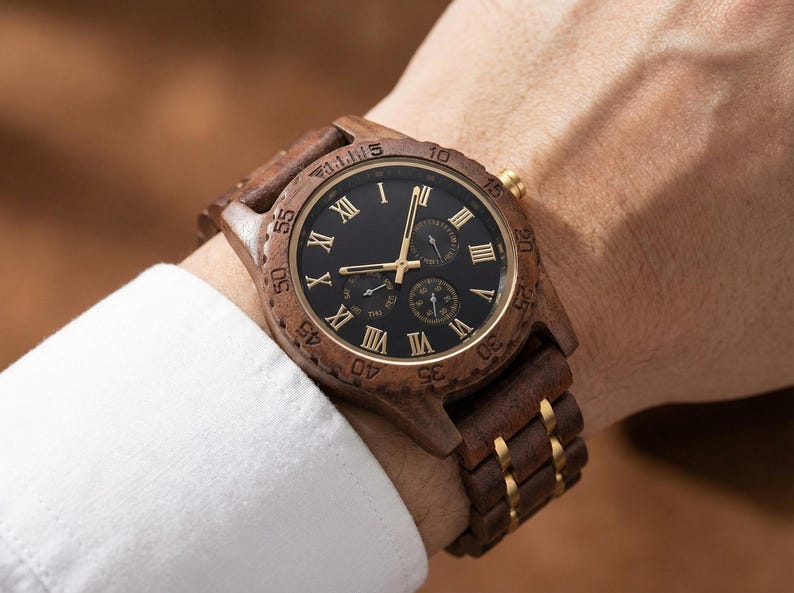 Peut inclure: Montre-bracelet en bois avec un cadran noir et des chiffres romains dor&eacute;s. La montre a une lunette et un bracelet en bois marron avec des accents dor&eacute;s. La montre est sur un poignet avec une manche de chemise blanche.