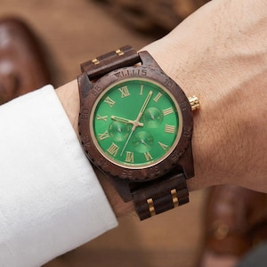Peut inclure: Une montre-bracelet en bois avec un cadran vert et des accents dor&eacute;s. La montre a des chiffres romains pour les heures et trois sous-cadrans. Le bracelet et le bo&icirc;tier sont en bois brun fonc&eacute;. La montre est sur un poignet.