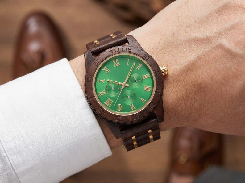 Peut inclure: Une montre-bracelet en bois avec un cadran vert et des accents dor&eacute;s. La montre a des chiffres romains pour les heures et trois sous-cadrans. Le bracelet et le bo&icirc;tier sont en bois brun fonc&eacute;. La montre est sur un poignet.
