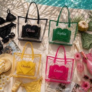 May include: Four clear tote bags with matching coloured handles and trim. Each bag contains a heart-shaped pouch featuring a name: Olivia in black, Sophia in green, Charlotte in yellow, and Emma in pink. The bags are arranged with various beach accessories.