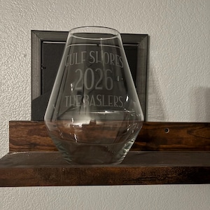 Puede incluir: Un jarrón de vidrio transparente con un cuerpo ancho y angulado. El jarrón tiene texto grabado que dice "GULF SHORES 2026 THE BASLERS". El jarrón se exhibe en un estante de madera oscura, con una imagen enmarcada en el fondo.