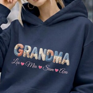 Könnte beinhalten: Marineblauer Kapuzenpullover mit dem Wort "GRANDMA" in bunten, gemusterten Buchstaben. Darunter stehen die Namen Lea, Mia, Sam und Ava, jeweils mit einem Herzsymbol. Der Pullover hat eine Vordertasche.