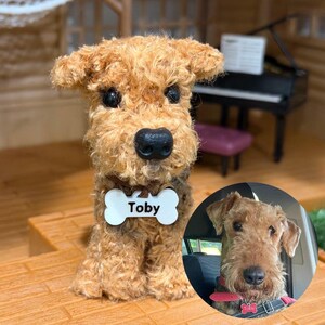 May include: A tan, curly-haired plush dog figurine with a black nose and dark eyes. A white, bone-shaped tag hangs from its neck, featuring the name Toby in black text. An inset circular photo shows a real dog with similar features wearing a collar.