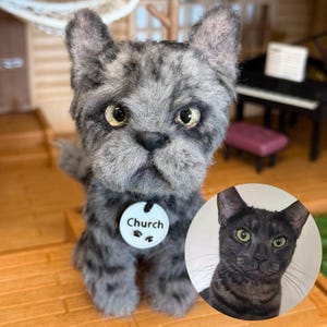 May include: A needle-felted grey tabby cat figurine with textured fur, pointed ears, and yellow eyes. The cat wears a white circular tag around its neck with the text Church and two black paw prints. A small circular photo of a real cat is included.