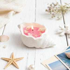 May include: A lit pink candle in a white seashell-shaped holder. The candle is decorated with small seashells and a starfish. The candle sits on a white wooden surface, with additional seashells and a starfish nearby.