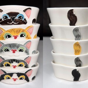 May include: A stack of white ceramic cat bowls. The bowls on the left feature painted cat faces in various colours and patterns. The bowls on the right have painted cat tails. A fun and whimsical design for cat lovers.