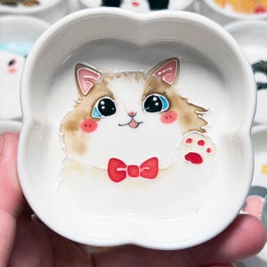 May include: A white ceramic dish in a four-leaf clover shape, decorated with a cartoon cat. The cat has a red bow tie, pink cheeks, and a waving paw. The cat's fur is brown and white, with large blue eyes.