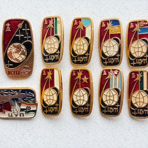 May include: A collection of ten rectangular metal enamel pins featuring space-themed designs with a globe, stars, and the Cyrillic text ЦУП. The pins display various national flag colors, including red, white, blue, green, and yellow, with gold-toned borders.