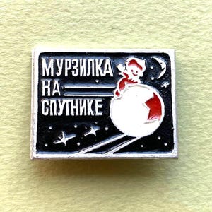 Puede incluir: Un pin rectangular de metal con fondo negro que muestra un satélite blanco, una figura roja y estrellas. El texto "МУРЗИЛКА НА СПУТНИКЕ" aparece en letras cirílicas blancas. El diseño incluye una luna creciente y líneas finas que representan movimiento.