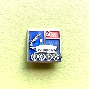 May include: A square metal pin featuring a blue background with a white lunar rover graphic. The rover has eight wheels, an antenna, and the text LUNOKHOD-1 in Cyrillic. A small red rectangle with a white hammer and sickle symbol is in the top right corner.