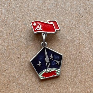 May include: A metal pin featuring a red flag with a white hammer and sickle symbol attached to a pentagonal pendant. The pendant displays a dark blue space scene with a silver rocket, three stars, and a green and red globe at the bottom.