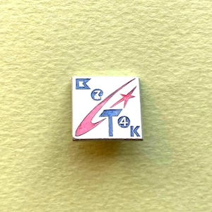 May include: A square metal pin with a white background featuring a blue letter B, a blue letter T, a blue letter K, and a blue number 4 inside a circle. A pink swooping shape crosses the design, accompanied by a red four-pointed star graphic.