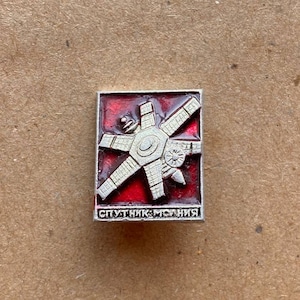 Puede incluir: Un pin metálico rectangular que presenta un diseño de satélite en tono plateado sobre un fondo de esmalte rojo intenso. El satélite tiene un cuerpo central con varios paneles extendidos. En la parte inferior, el texto "СПУТНИК-МОЛНИЯ" está grabado en plata.