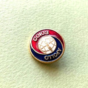 May include: A circular metal pin featuring a red and blue enamel design with a central white globe graphic. The text "СОЮЗ" is written in white Cyrillic letters on the red section, and the word "APOLLO" is written in white on the blue section.