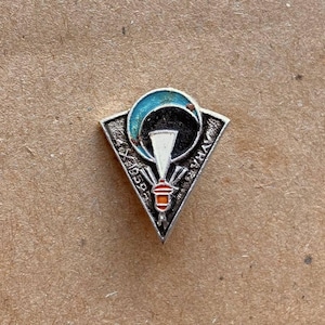 Può includere: Una spilla triangolare in metallo color argento con un grafico centrale a paracadute bianco, una forma a mezzaluna blu e un piccolo dettaglio arancione. La spilla include il testo 1959 e caratteri cirillici sulla superficie argentata.