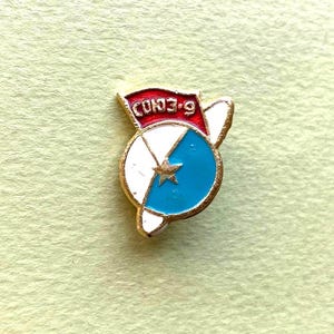 May include: A gold-toned metal lapel pin featuring a circular design with a white and light blue split background, a central gold star, and a red banner at the top with the Cyrillic text СОЮЗ-9 in gold lettering, accented by white wing-like shapes.