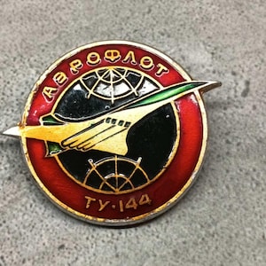 Pode incluir: Um pin vintage da Aeroflot com uma borda vermelha e dourada. O pin apresenta um avião estilizado em amarelo e verde sobre um fundo preto, com a palavra cirílica "AEROFOT" e "TU-144" em letras douradas. O pin é circular.