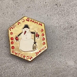 Può includere: Spilla esagonale color oro con un pupazzo di neve bianco con cappello nero, guanti rossi e una scopa. La spilla ha un testo rosso in un motivo circolare attorno al pupazzo di neve. Il testo recita "Festa invernale russa."
