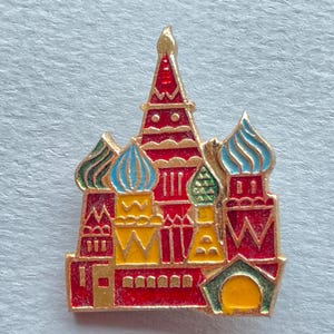 May include: A colorful enamel pin depicting Saint Basil's Cathedral. The pin features red, gold, yellow, blue, and green architectural details. The pin is a miniature representation of the iconic Russian landmark.
