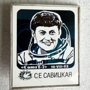 May include: A square metal pin featuring a black and white portrait of a smiling astronaut in a space suit. The text reads Soyuz T-7, 19-VIII-82, and S.E. Savitskaya, along with a circular logo containing a bird and the Cyrillic text CCCP KOSMOS.