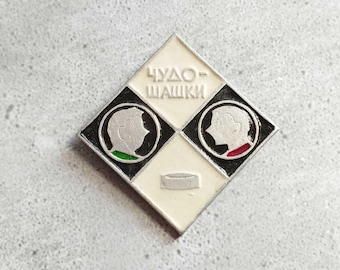 Puede incluir: Un pin vintage con forma de diamante, con el texto cirílico "ЧУДО-ШАШКИ". Presenta dos cuadrados en blanco y negro con siluetas de perfil, uno con acentos verdes y otro con acentos rojos. Un pequeño rectángulo horizontal blanco está en la parte inferior.
