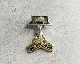 May include: A silver-toned pin featuring a hockey net above a pair of ice skates. The skates are detailed with laces and blades. The pin is likely made of a metal alloy and has a vintage appearance.