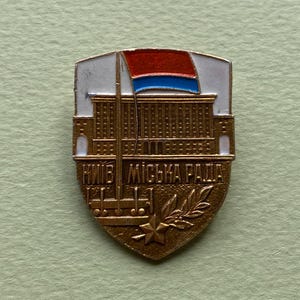 May include: A shield-shaped metal pin featuring a gold-toned building with a flagpole and a red and blue flag. The text "КИЇВ МІСЬКА РАДА" is embossed at the bottom, accompanied by a small star and a laurel branch design on a white background.