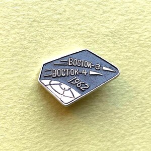 May include: A silver-toned metal pin shaped like a rhombus with a dark gray center. The pin features the text "BOCTOK-3", "BOCTOK-4", and "1962" in raised silver lettering, along with a stylized graphic of a globe grid in the lower corner.