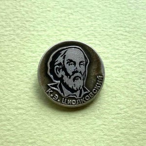 Puede incluir: Un broche circular de metal plateado con un retrato en relieve de un hombre con barba. El texto cirílico K.Э. ЦИОЛКОВСКИЙ está grabado a lo largo del borde inferior del broche, que presenta un acabado oscuro y oxidado en las zonas hundidas.